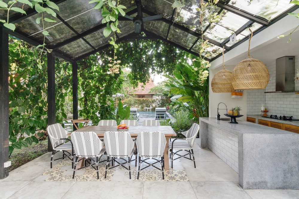 Open-air kitchen with empty table and chairs, set against a backdrop of fresh green plants, highlighting the role of landscape service in outdoor living spaces.